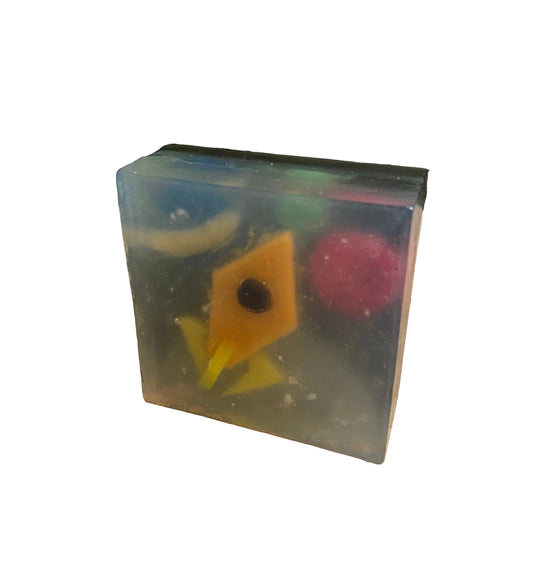A translucent rectangular resin block or candle base on a wooden surface, containing colorful embedded shapes including a yellow diamond, pink oval, and blue curved piece, with a kitchen countertop and tiled backsplash in the background.